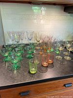 Wide view of various colorful stemmed glasses and tumblers arranged on countertop including green dessert glasses, yellow and amber stemware, and orange tumblers.