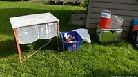 Lot items arranged outside on grass next to siding, showing cooler table, blue plastic bin, Coleman cooler, and Rubbermaid drink dispenser.