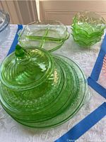 Overview of stacked green depression glass dinner plates, lidded round serving bowl, divided octagonal dish, and several ruffled edge small bowls.