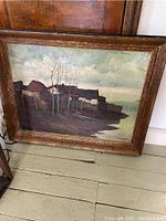 Full frontal view of the framed riverside village oil painting showing brown-gold frame, buildings, bare trees, and water reflections.