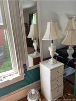 Framed mirror and white nightstand with drawer shown near window with white lamp on top. Room interior and outside visible through window.