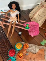 Pocahontas doll seated on pink fabric leaf with raccoon figurine, toy food items, and beige teepee tent in background.