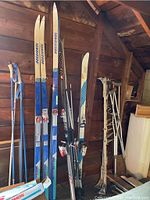 Photo showing cross country and downhill skis standing against wooden wall, along with ski poles