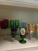 A shelf with 3 sets of colorful etched stemmed glasses in red, green, blue, amber, and a single decorative green glass with floral medallion.