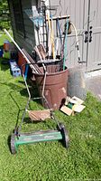 Front view of lot showing Scotts Classic green push mower in front, brown plastic waste can behind filled with garden tools including metal rakes, poles, and clippers, and rolled chicken wire to the side.