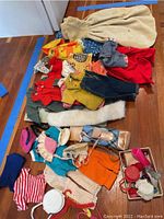 Photo showing spread of vintage doll clothes including dresses, pants, shirts, shoes, and hats on wooden floor with cream blanket and plastic hangers.