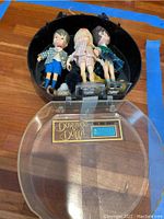 Three vintage Dolly Darling dolls in a black circular plastic case with clear lid, laid open revealing dolls inside and label on base of the case.