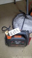 Front angled photo of the MotoMaster battery charger with attached cables and carrying bag visible, showing overall condition and size.