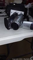 Front side view of silver Canon HDV video camera on table with black carrying case and user manual behind it.