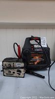 Photo showing MotoMaster Eliminator mobile booster pack with cables and carrying handle alongside the Texson AC/DC variable rate auto charger.