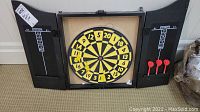 Open black Churchill dart board wooden case showing yellow and black dart board with numeric segments. Left door has cricket game scoreboard, right door has scoring board and 3 red darts held in place.
