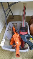 Three garden hand tools including a black and orange/red hedge trimmer, shrubber, and trimmer grouped in a plastic basket, placed on carpeted floor.