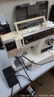 Side view of white Singer sewing machine showing carrying handle, stitch options panel, large dial, and attached foot pedal on table.