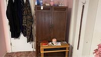View of a dark stained wooden armoire with two slatted doors, metal handles and lock. The armoire is placed against a wall with some items on top and in front.
