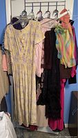 Closeup image showing various ladies' dresses (yellow floral, pink satin, black lace) and a colorful scarf hanging on hangers