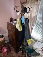 Tall dark wooden coat rack with multiple curved hooks holding clothing items and a mask, showing surface wear and scratches; placed beside a dresser in a corner by a window with curtains.