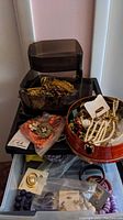 Overview photo showing plastic containers and a round red dish filled with assorted costume jewelry including bracelets, pins, and earrings, some with coral beads and purple tones.