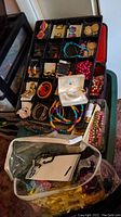 Overview of costume jewelry organized in a black tray with compartments, including earrings, pins, necklaces, hairbands, and wraps, plus additional loose pieces in plastic bags underneath.
