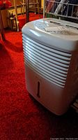 Full view of the white portable dehumidifier on red carpet, showing front grille and water level window.