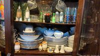 Wooden cabinet shelf with stacked white plates, cups, and saucers featuring blue floral metallic embellishments along with salt and pepper shakers and small condiment bottles
