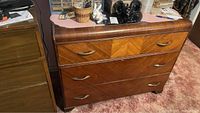Front angle shot of vintage wooden dresser showing three drawers with chevron veneer pattern and curved metal handles, placed on carpeted floor.