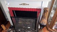 Full view of white faux mantle with red tile-like trim inside, black fireplace screen in front, and a carved wooden sculpture on the right.