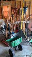 Multiple wooden-handled garden tools including rakes, shovels, and a push broom arranged vertically against a rack, plus a green Scotts SpeedyGreen 1000 seed spreader in the foreground.