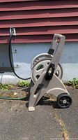 Side view of the beige plastic hose reel with attached green hose, black crank and wheels, placed on a paved outdoor surface against a red-sided house