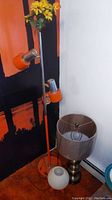Full view of metal floor lamp with orange details next to brushed metal table lamp with dark shade and plastic desk lamp shade on floor.