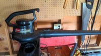 Black Toro Super Blower Vac with long nozzle and handle positioned on a wooden surface by a wall outlet and pegboard.