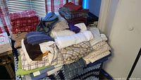 Wide view of assorted folded linens, towels, and clothing piled on a surface near a window with blinds.
