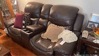 Photo showing front view of dark brown leather love seat with two seats, padded high backs, and a center fold-down armrest with two cupholders. Cushions and throws placed on seat surfaces.