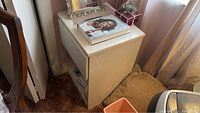Wooden side table showing simple rectangular shape with a lower shelf and some surface wear. Top surface holding some books and decorative items.
