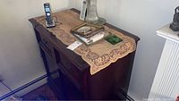 Wooden dark-stained sideboard with lace runner, cordless phone, and glass items on top, showing front and top surfaces at an angle.