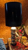 Photo showing a lit table lamp with four stacked chrome balls and large black cylindrical shade on a glass surface with surrounding decorative items.