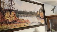 Close-up image of a large framed wall art print depicting a river and forest scene with autumn foliage and mountains in the distance.