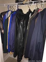 Photo showing a row of jackets and coats hanging on hangers inside a closet, including a black leather jacket and several blue coats.