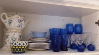 Photo displaying all dishware and glassware items stacked and lined up in cabinet. Shows teapot with floral blue design and candle warmer, various plates and bowls in blue and white, multiple blue drinking glasses, and clear martini glasses with blue stems.