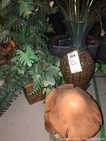 Image shows multiple faux plants including a tall one in a woven wicker pot with metal stand, smaller plants in square wicker pots and a decorative brown mushroom.