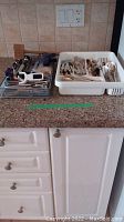 Two trays on a kitchen countertop; one white plastic tray with an assortment of flatware including spoons, forks, butter knives, and other utensils; a smaller metal tray holding additional kitchen utensils such as tongs, spatula, pie lifter, and scissors.