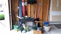 Wide view of garage containing black woven planter table with faux plants, wooden planter boxes on floor, broom and other garden tools hung on wall, and watering pail on floor.