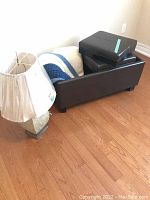 Table lamp, two knitted blankets, and dark brown faux leather ottoman with removable cushions on wooden floor near wall.