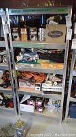 Metal shelving with assorted paint cans, power bars, rollers, cords, hand tools, and hardware items arranged on four shelves.