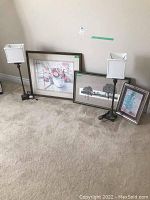 Overall photo showing the three framed prints arranged on floor leaning against wall and two tall matching lamps with white square shades and dark bases on carpeted floor