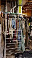 Photo of a metal rolling clothing rack loaded with various hanging linens like tablecloths, runners, and curtains in different colors and patterns. Visible wooden ceiling and basement setting.