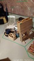 Photo showing child-size white wooden rocking chair, two wooden TV tables in a folded position, multiple baskets and tins placed on basement floor.