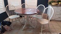 Wood round dining table with four white wood chairs around it, set on tiled floor near kitchen cabinets.