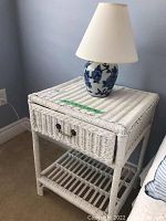 Front angled view of white wicker side table with blue and white floral ceramic lamp on top