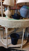 Photo showing off-white wicker half-moon table with two lamps on top and two black baskets, one on the table and one on the lower shelf.