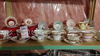 Eight teacups and saucers displayed on a metal shelf against plastic backing.